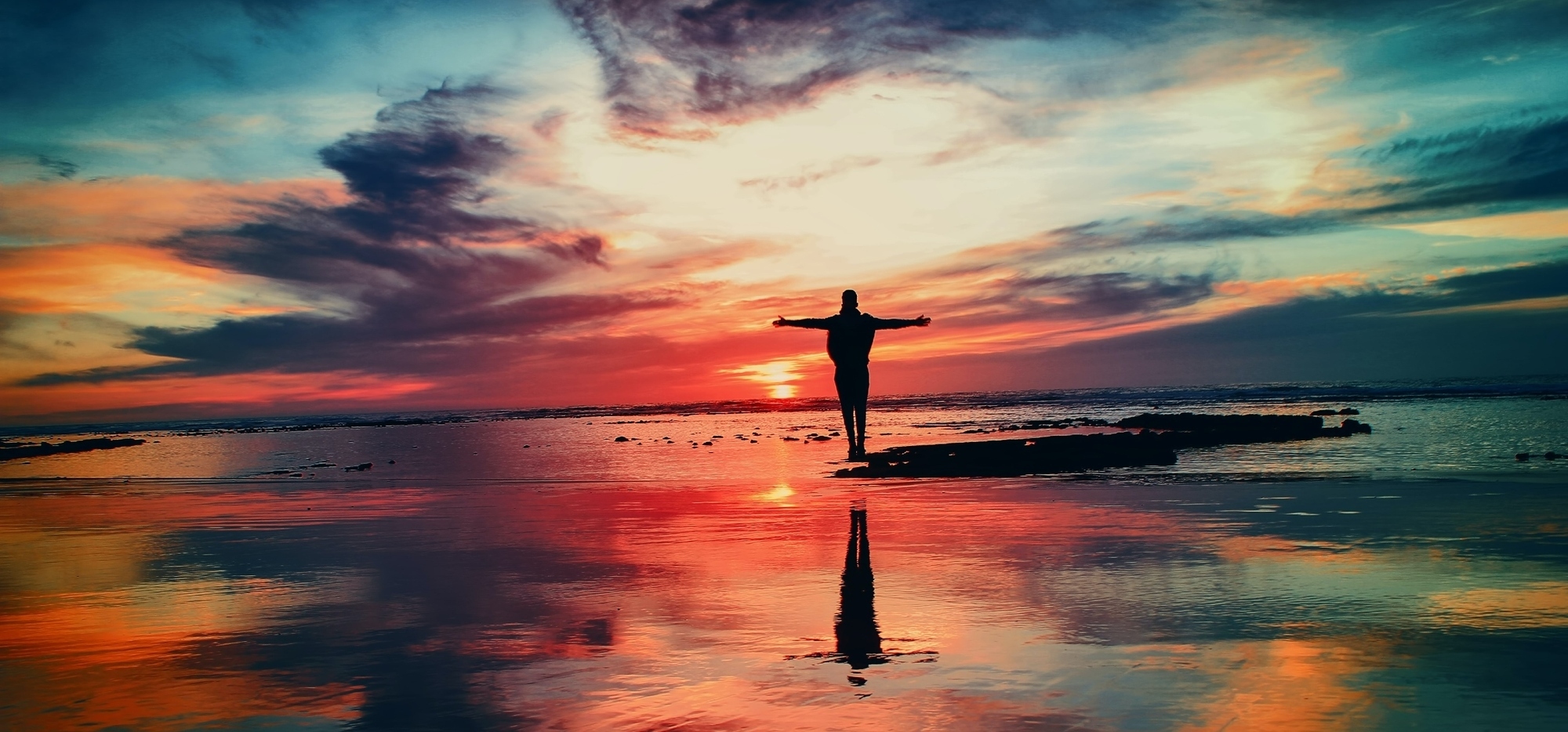 Man on beach arms outstretched facing a colourful sunset